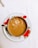 A porcelain cup filled with milk tea sits on a matching saucer featuring red floral designs. A splash of milk is captured mid-air, creating ripples and bubbles in the tea. The setting is on a white tablecloth.