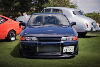 A sleek blue sports car is prominently positioned in the foreground, with its distinct headlights and license plate visible. Behind it, an orange vintage car with noticeable decals and a shiny silver classic car are parked on green grass. Trees provide a natural backdrop under a partly cloudy sky.