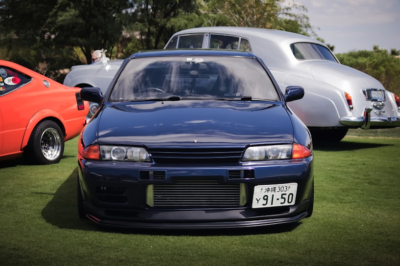 A sleek blue sports car is prominently positioned in the foreground, with its distinct headlights and license plate visible. Behind it, an orange vintage car with noticeable decals and a shiny silver classic car are parked on green grass. Trees provide a natural backdrop under a partly cloudy sky.