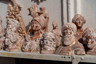 A collection of detailed, classical-style terracotta busts and sculptures are arranged on a shelf. The busts depict various historical or mythical figures with intricate details such as helmets, hairstyles, and facial expressions. The sculptures have a weathered and rustic appearance, suggesting they may be replicas of ancient works.
