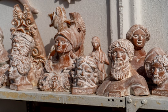 A collection of detailed, classical-style terracotta busts and sculptures are arranged on a shelf. The busts depict various historical or mythical figures with intricate details such as helmets, hairstyles, and facial expressions. The sculptures have a weathered and rustic appearance, suggesting they may be replicas of ancient works.