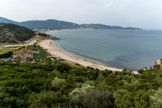 A coastal landscape featuring a serene beachfront with gentle waves lapping against the shore. The foreground is lush with various green vegetation leading to a quaint house with a terracotta roof and palm trees nearby. A winding road runs along the coast, lined with a few parked vehicles. The background showcases distant hills and a small town across the bay, under a slightly overcast sky.