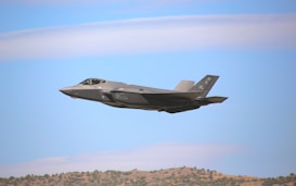 A military jet, specifically a stealth fighter, flies across a clear blue sky with a few wispy clouds. The aircraft is grey, with distinct sharp angles and an aerodynamic design. Below, a landscape with low hills and sparse vegetation is visible.