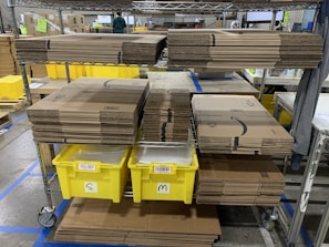Stacks of cardboard boxes are neatly organized on a metal shelving unit with yellow plastic bins labeled 'S' and 'M' below them. The setting appears to be a warehouse or storage area with visible industrial flooring and safety line markings.