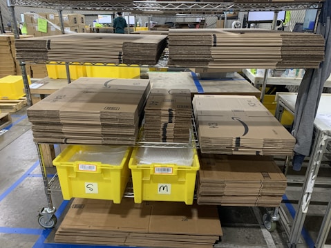 Stacks of cardboard boxes are neatly organized on a metal shelving unit with yellow plastic bins labeled 'S' and 'M' below them. The setting appears to be a warehouse or storage area with visible industrial flooring and safety line markings.