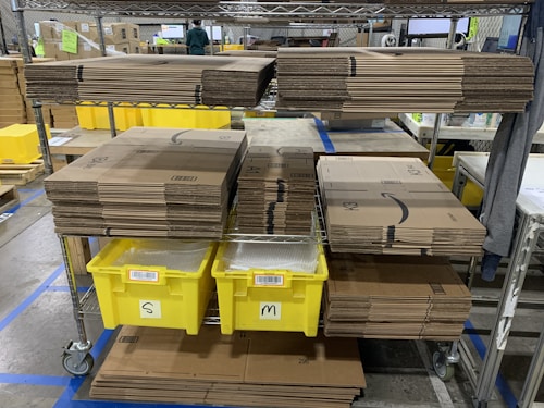 Stacks of cardboard boxes are neatly organized on a metal shelving unit with yellow plastic bins labeled 'S' and 'M' below them. The setting appears to be a warehouse or storage area with visible industrial flooring and safety line markings.