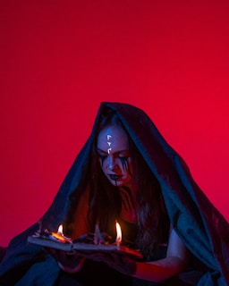 A person cloaked in dark fabric sits against a vivid red background, holding an open book with flames rising from its pages. Black makeup accents their eyes and cheeks, creating a dramatic and mysterious appearance. Symbols are painted on their forehead.