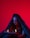A person cloaked in dark fabric sits against a vivid red background, holding an open book with flames rising from its pages. Black makeup accents their eyes and cheeks, creating a dramatic and mysterious appearance. Symbols are painted on their forehead.