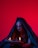 A person cloaked in dark fabric sits against a vivid red background, holding an open book with flames rising from its pages. Black makeup accents their eyes and cheeks, creating a dramatic and mysterious appearance. Symbols are painted on their forehead.