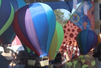 A collection of colorful, miniature hot air balloons made of fabric, suspended in the air at an outdoor event. The balloons feature a variety of colors, including blue, purple, green, and red. In the background, there is a vibrant, patterned banner with bold designs and an abstract collage of shapes, along with a subtle crowd of people.