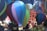 A collection of colorful, miniature hot air balloons made of fabric, suspended in the air at an outdoor event. The balloons feature a variety of colors, including blue, purple, green, and red. In the background, there is a vibrant, patterned banner with bold designs and an abstract collage of shapes, along with a subtle crowd of people.