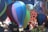 A collection of colorful, miniature hot air balloons made of fabric, suspended in the air at an outdoor event. The balloons feature a variety of colors, including blue, purple, green, and red. In the background, there is a vibrant, patterned banner with bold designs and an abstract collage of shapes, along with a subtle crowd of people.