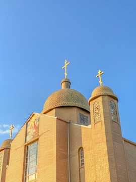 A brick building features ornate architecture with domes and crosses on top. Sunlight casts warm shadows on the structure, adding depth to the intricate design. A large, colorful mural is displayed above a series of windows, enhancing the religious theme.