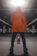 A person in an orange hoodie with the text 'ARE WE HAVING FUN YET?' stands in an empty, dimly lit parking garage. They hold a knife in one hand and face a small doll dressed in colorful overalls, resembling a toy character known for being creepy.