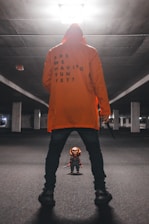 A person in an orange hoodie with the text 'ARE WE HAVING FUN YET?' stands in an empty, dimly lit parking garage. They hold a knife in one hand and face a small doll dressed in colorful overalls, resembling a toy character known for being creepy.