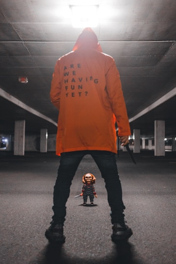 A person in an orange hoodie with the text 'ARE WE HAVING FUN YET?' stands in an empty, dimly lit parking garage. They hold a knife in one hand and face a small doll dressed in colorful overalls, resembling a toy character known for being creepy.