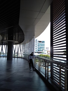A modern architectural walkway with large windows allowing plenty of natural light. A person is leaning casually against the glass railing, gazing outside. The design of the area is sleek, featuring a high ceiling with unique patterns and a tiled floor. The view outside includes a glass building and lush greenery.