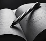 An open book with a pen resting diagonally across the pages. The text is slightly blurred, and the overall tone is in black and white, creating a focused and intimate atmosphere. The pen casts a shadow on the page, enhancing the depth and lighting contrast.
