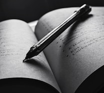 An open book with a pen resting diagonally across the pages. The text is slightly blurred, and the overall tone is in black and white, creating a focused and intimate atmosphere. The pen casts a shadow on the page, enhancing the depth and lighting contrast.