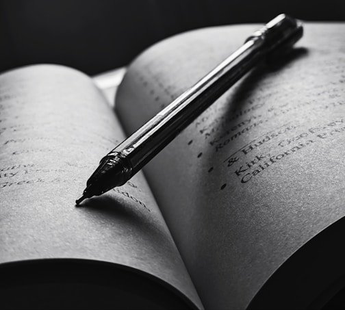 An open book with a pen resting diagonally across the pages. The text is slightly blurred, and the overall tone is in black and white, creating a focused and intimate atmosphere. The pen casts a shadow on the page, enhancing the depth and lighting contrast.