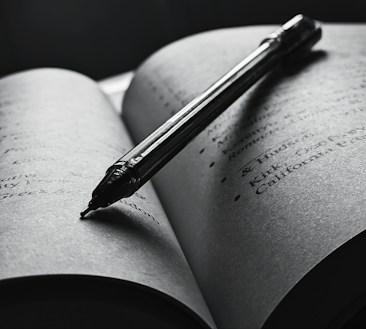 An open book with a pen resting diagonally across the pages. The text is slightly blurred, and the overall tone is in black and white, creating a focused and intimate atmosphere. The pen casts a shadow on the page, enhancing the depth and lighting contrast.