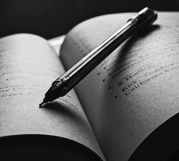 An open book with a pen resting diagonally across the pages. The text is slightly blurred, and the overall tone is in black and white, creating a focused and intimate atmosphere. The pen casts a shadow on the page, enhancing the depth and lighting contrast.