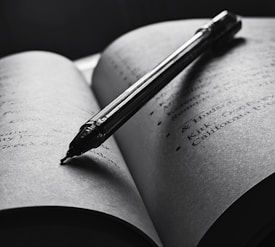 An open book with a pen resting diagonally across the pages. The text is slightly blurred, and the overall tone is in black and white, creating a focused and intimate atmosphere. The pen casts a shadow on the page, enhancing the depth and lighting contrast.