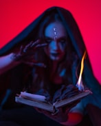 A mysterious figure cloaked in dark fabric reaches towards an old, open book with one hand. The book appears to be burning, with flames emanating from its pages. Strange symbols glow on the figure's forehead, and the lighting casts an eerie, otherworldly glow.