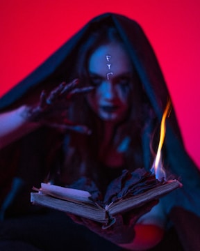 A mysterious figure cloaked in dark fabric reaches towards an old, open book with one hand. The book appears to be burning, with flames emanating from its pages. Strange symbols glow on the figure's forehead, and the lighting casts an eerie, otherworldly glow.