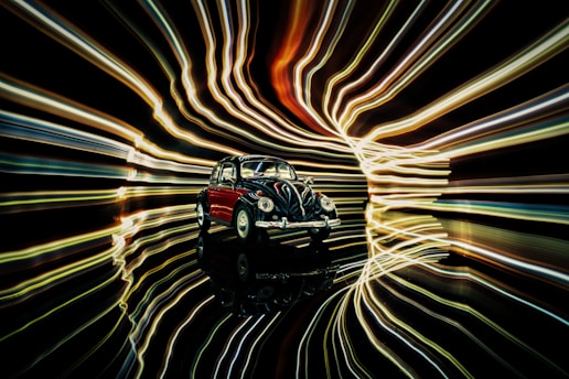 A classic, vintage car model is positioned against a backdrop of dynamic, swirling light trails. The lights create a sense of motion and energy, contrasting with the stationary car. The reflective surface beneath the car enhances the illusion of depth and movement.