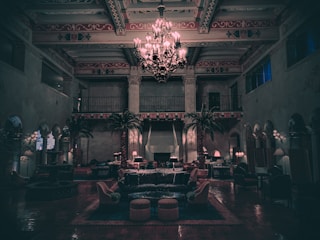 The image features an elegant, dimly lit interior of a large room with high ceilings adorned with intricate designs. In the center, a grand chandelier emits a soft glow, illuminating the surrounding space filled with plush seating arrangements and tall palm plants. The floor is covered in rich-looking tiles, while the walls display an ornate decorative pattern with arches and balconies.
