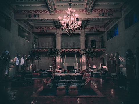 The image features an elegant, dimly lit interior of a large room with high ceilings adorned with intricate designs. In the center, a grand chandelier emits a soft glow, illuminating the surrounding space filled with plush seating arrangements and tall palm plants. The floor is covered in rich-looking tiles, while the walls display an ornate decorative pattern with arches and balconies.