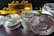 A stack of silver-colored cryptocurrency coins is prominently displayed in front of several shiny gold bars. The coins feature intricate engraving and digital circuit designs.