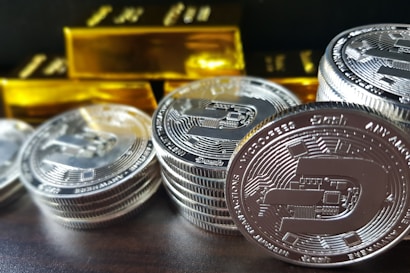 A stack of silver-colored cryptocurrency coins is prominently displayed in front of several shiny gold bars. The coins feature intricate engraving and digital circuit designs.