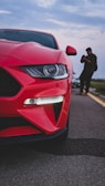 A close-up of the front end of a red sports car is captured on a road, with its sleek design and prominent headlight in focus. In the background, a person is seen taking photos or videos with a mobile phone.