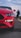 A close-up of the front end of a red sports car is captured on a road, with its sleek design and prominent headlight in focus. In the background, a person is seen taking photos or videos with a mobile phone.