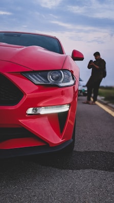 A close-up of the front end of a red sports car is captured on a road, with its sleek design and prominent headlight in focus. In the background, a person is seen taking photos or videos with a mobile phone.