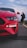 A close-up of the front end of a red sports car is captured on a road, with its sleek design and prominent headlight in focus. In the background, a person is seen taking photos or videos with a mobile phone.