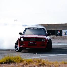 A red car performs a drift maneuver on a race track, with smoke billowing from its tires. The foreground is slightly blurred with some grass peeking in, and the background is an open, barren landscape with a hint of shrubbery. The car is angled towards the camera, emphasizing its dynamic movement.
