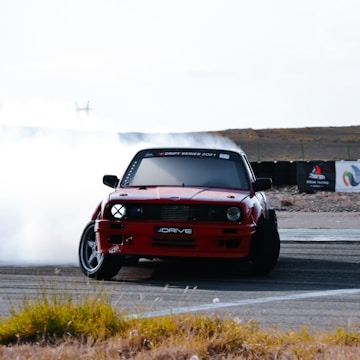 A red car performs a drift maneuver on a race track, with smoke billowing from its tires. The foreground is slightly blurred with some grass peeking in, and the background is an open, barren landscape with a hint of shrubbery. The car is angled towards the camera, emphasizing its dynamic movement.