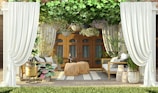 An outdoor seating area features elegant white curtains framing the space. The area is furnished with two wicker chairs and a table, surrounded by vibrant green plants and hanging floral baskets. The wooden doors in the background add a warm touch, while a striped rug adds texture to the floor.