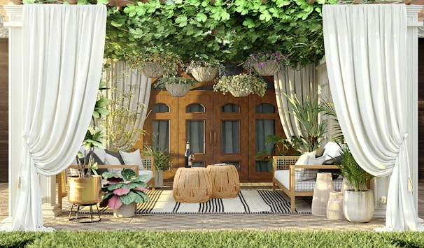 An outdoor seating area features elegant white curtains framing the space. The area is furnished with two wicker chairs and a table, surrounded by vibrant green plants and hanging floral baskets. The wooden doors in the background add a warm touch, while a striped rug adds texture to the floor.