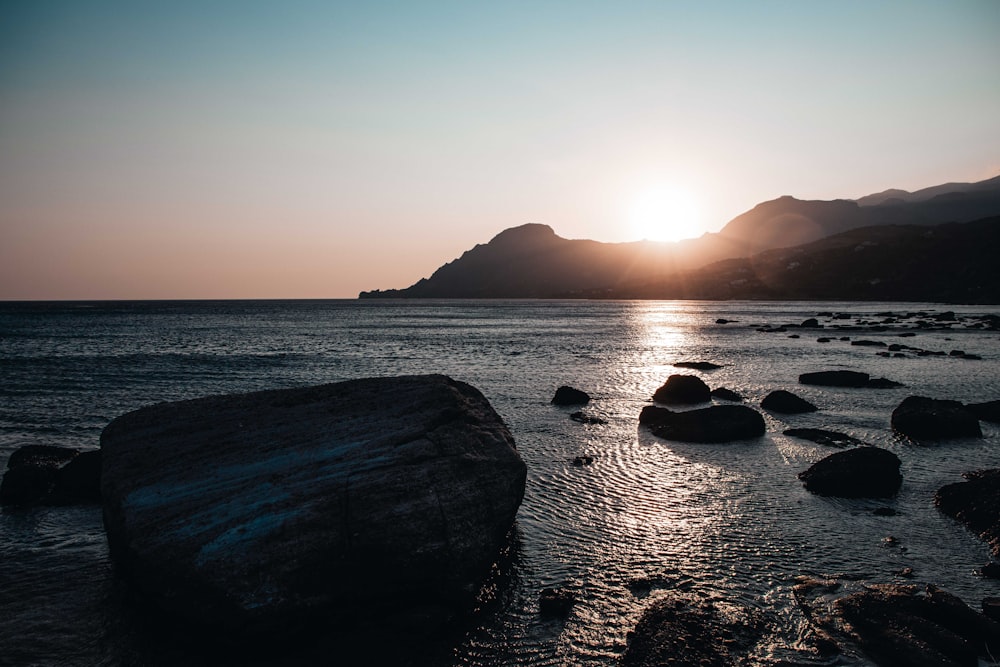 the sun is setting over the ocean with rocks in the foreground