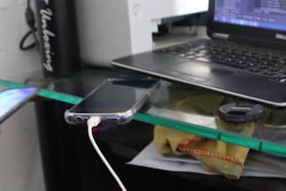 A workspace setup with a laptop on the right side, displaying a screen with code or text editor. A smartphone is charging with a cable connected to it, lying near the edge of a glass desk. A camera lens cap is also present on the desk, along with some papers partially visible beneath the glass surface. A thermos or tumbler with text on it is positioned to the left.