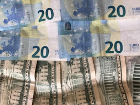 Several banknotes arranged in a slightly disorganized manner. The upper part displays four 20 Euro notes with a blue and yellow color scheme, featuring European architecture motifs. Below them, multiple 20 US dollar bills with green and white tones are visible, showing images of the White House.