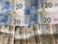 Several banknotes arranged in a slightly disorganized manner. The upper part displays four 20 Euro notes with a blue and yellow color scheme, featuring European architecture motifs. Below them, multiple 20 US dollar bills with green and white tones are visible, showing images of the White House.