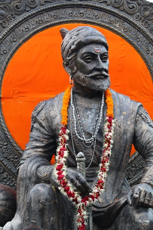 A majestic statue of a historical figure adorned with a garland of marigolds and red flowers. The statue, carved with intricate details, is seated prominently against a vibrant orange background. The figure wears a traditional headgear and multiple beaded necklaces, holding a sword.
