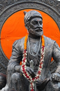 A majestic statue of a historical figure adorned with a garland of marigolds and red flowers. The statue, carved with intricate details, is seated prominently against a vibrant orange background. The figure wears a traditional headgear and multiple beaded necklaces, holding a sword.