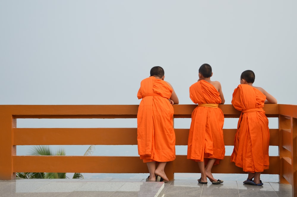 three people in orange robes standing on a bench