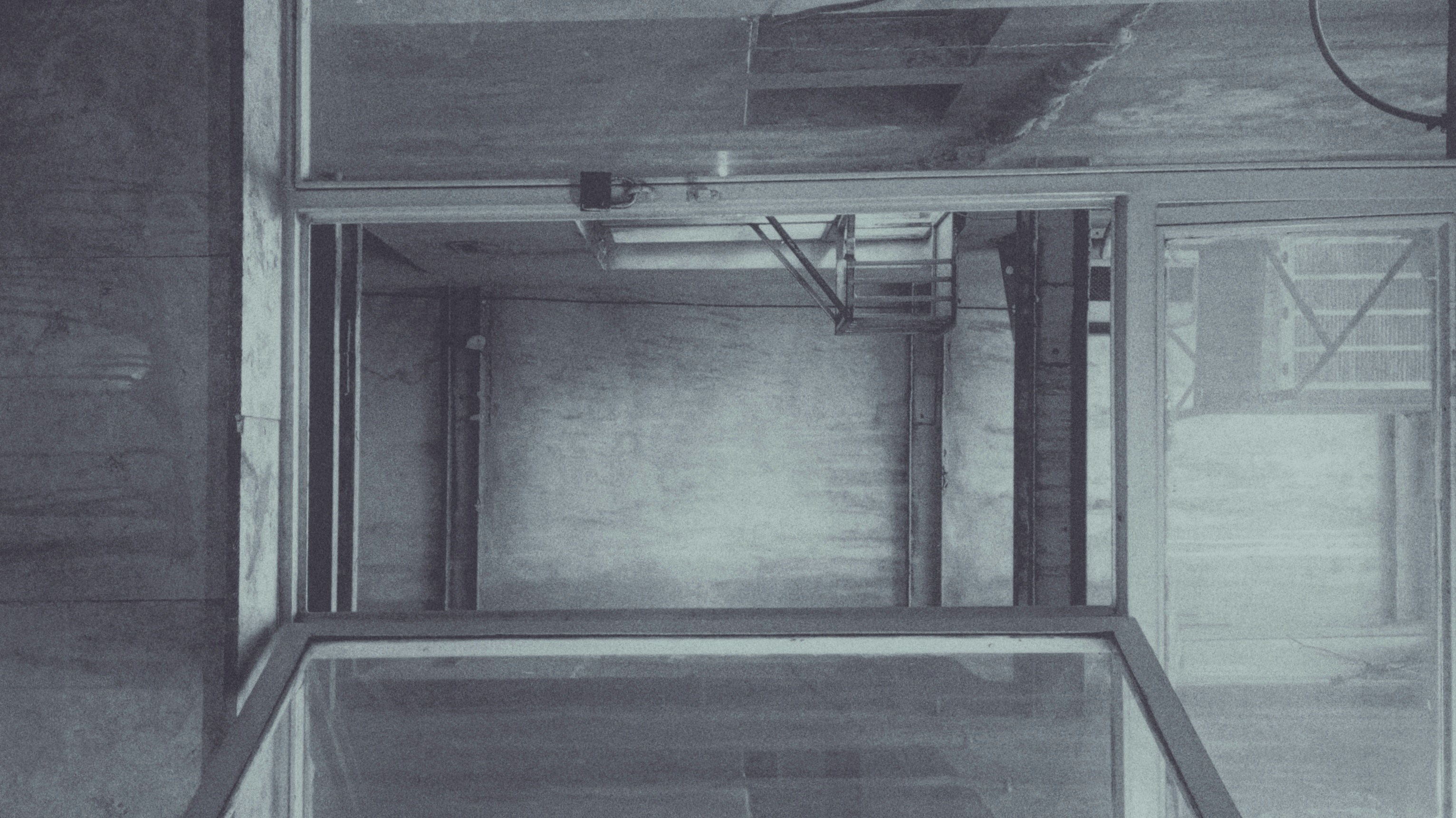 An empty, industrial elevator shaft with visible metal beams and concrete walls. The structure is framed by glass panels and has a utilitarian design. Subtle lighting casts soft shadows, adding depth to the interior.